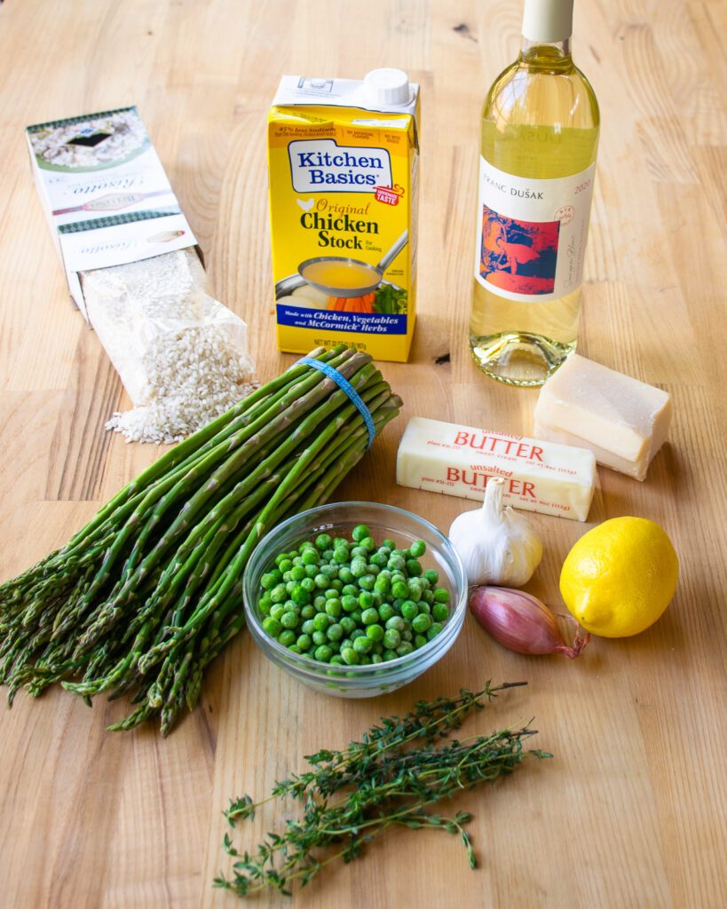 Ingredients on a butcher block counter - asparagus, peas, thyme, lemon, shallot, garlic, butter, parmesan cheese, stock, wine and rice.