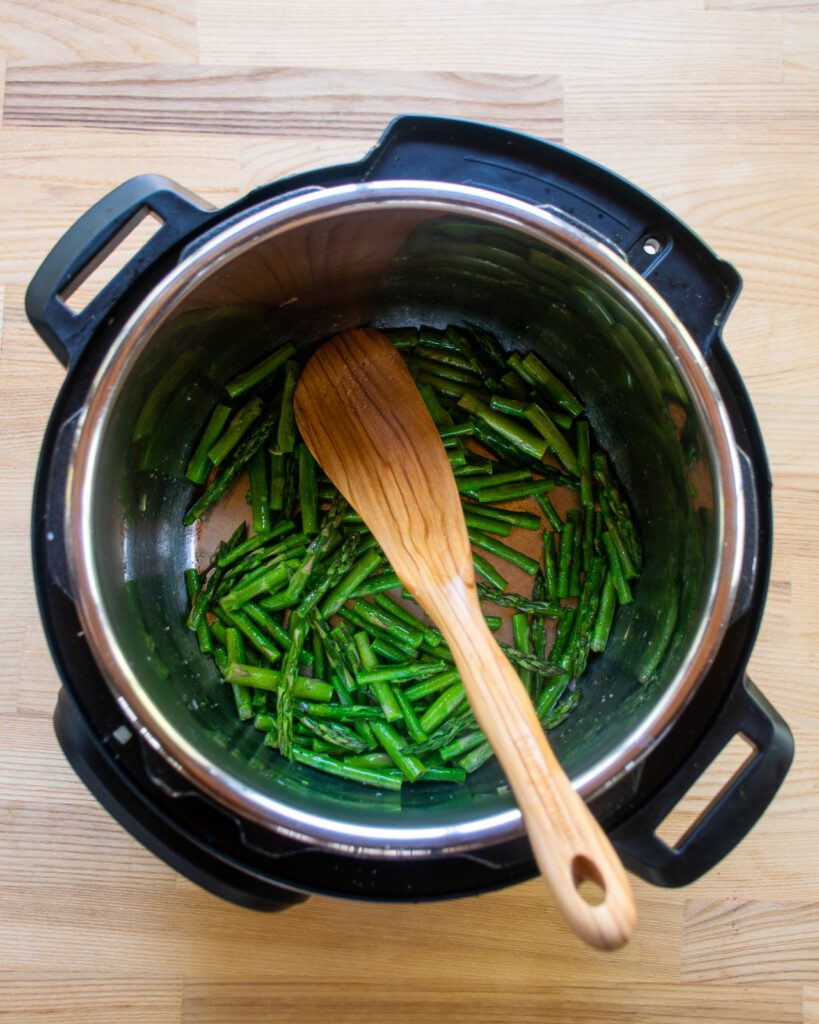 Looking down into a pressure cooker with asparagus sautéing and a wooden spoon sticking out.
