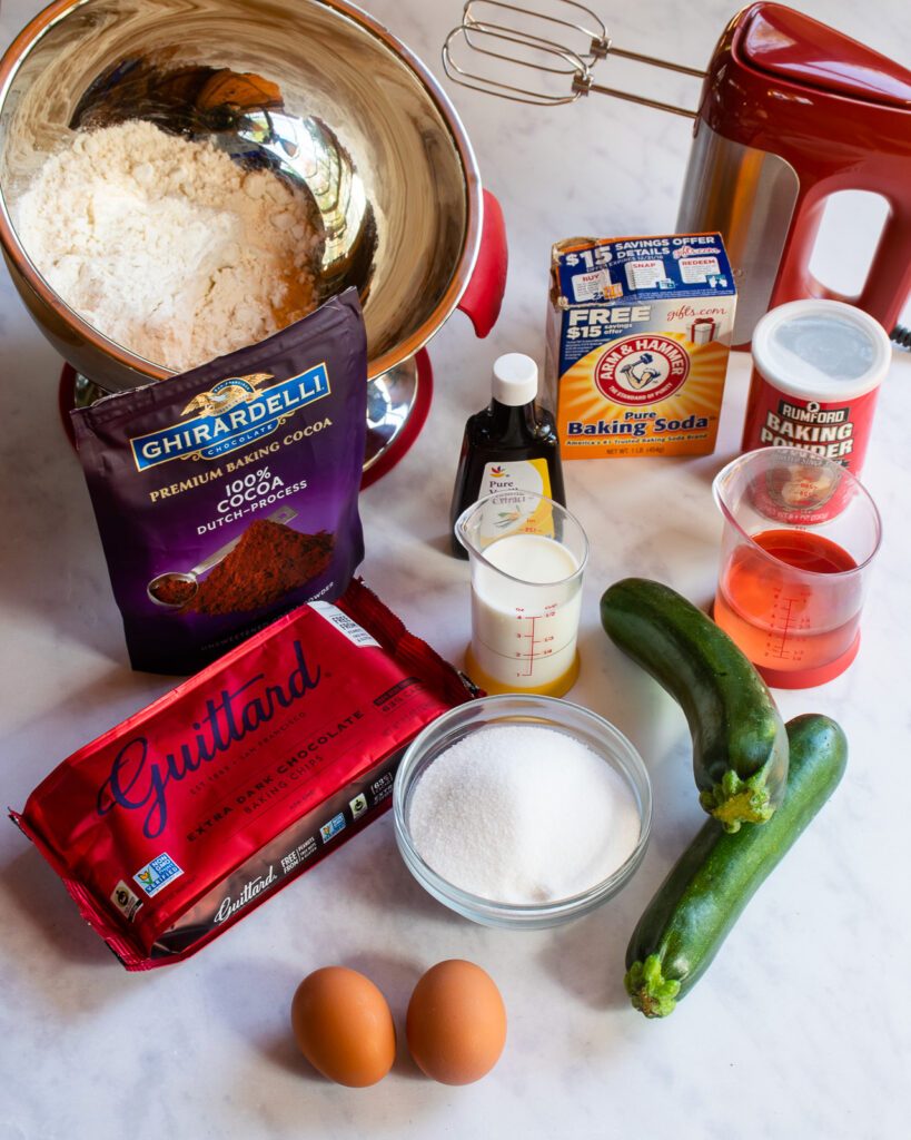 INgredients on a marble countertop - chocolate chips, sugar, eggs, zucchini, cocoa powder and other baking ingredients.
