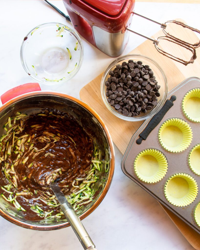Looking down on a mixing bowl with a spatula stirring grated zucchini into chocolate batter with a hand mixer and muffin pan nearby.