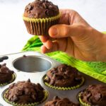 A hand holding a chocolate zucchini muffin above a muffin pan with a green towel in the background.
