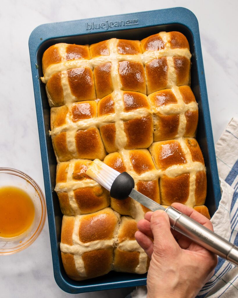 A hand brushing glaze onto cooked hot cross buns.