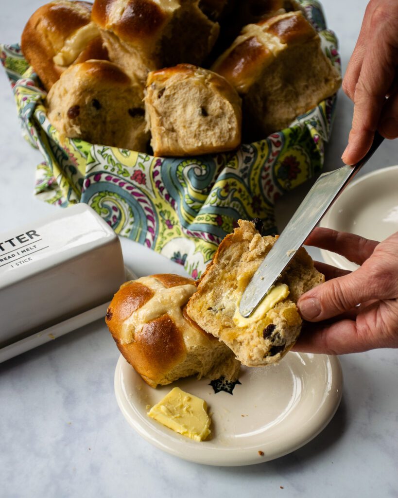 A hand brushing butter onto half of a hot cross bun with more in the background.