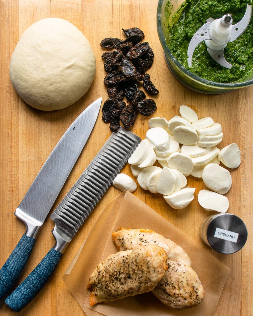 Ingredients on a cutting board with two knives - pizza dough, sun dried tomatoes, sliced fresh mozzarella, cooked chicken breast, oregano and basil.