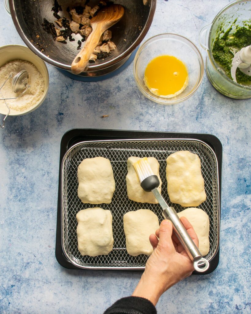 A hand brushing egg wash onto hot pockets in an air fryer basket.