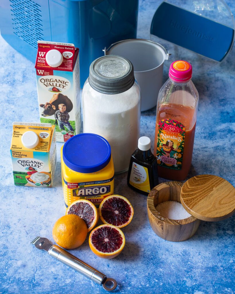 Ingredients on a blue counter - cream, milk, sugar, blood orange juice, cornstarch, salt, vanilla and some halved blood oranges.