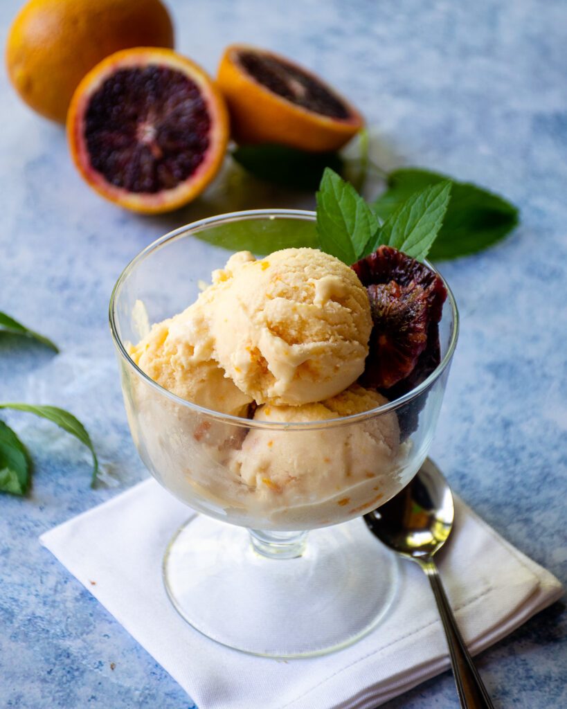 Blood orange gelato in a glass bowl with a halved blood orange and mint leaves around.