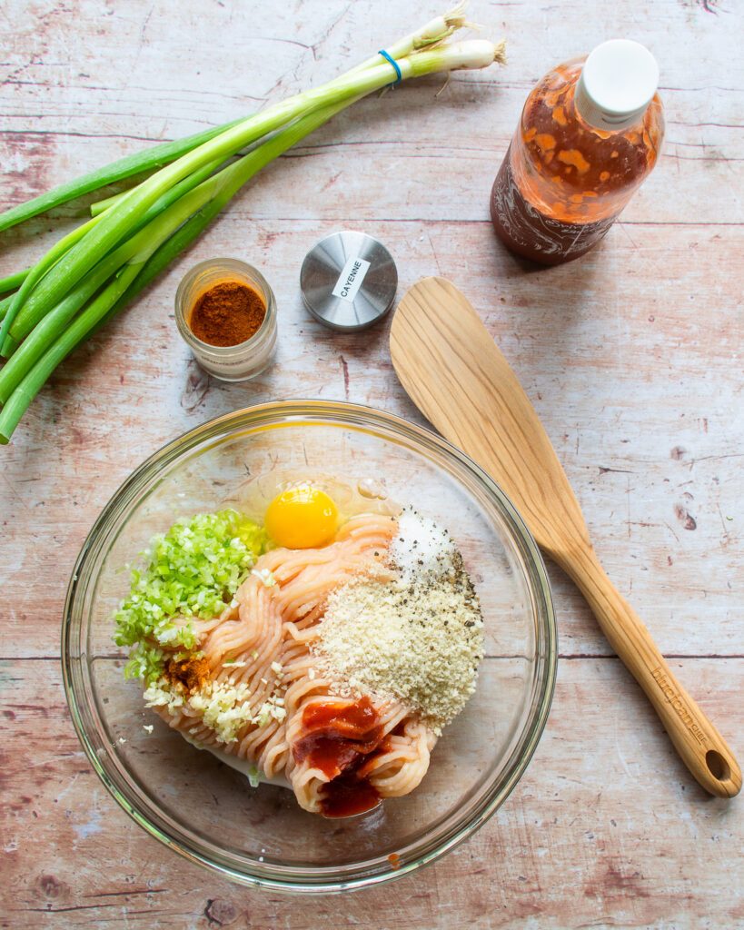 Ingredients in a bowl - ground chicken, scallions, spices, breadcrumbs, egg, with a wooden spoon and a bunch of scallions next to it.