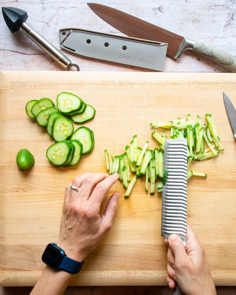 Hands cutting cucumber into sticks on a wooden cutting board.