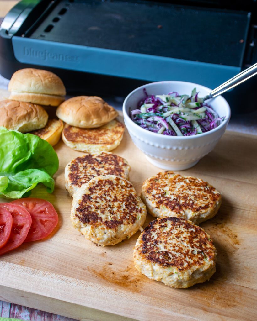 four zesty chicken burger patties on a wooden cutting board with cucumber slaw, tomato, lettuce and an electric grill in the background.