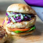 A chicken burger with cucumber slaw on a wooden cutting board with a purple napkin in the background.