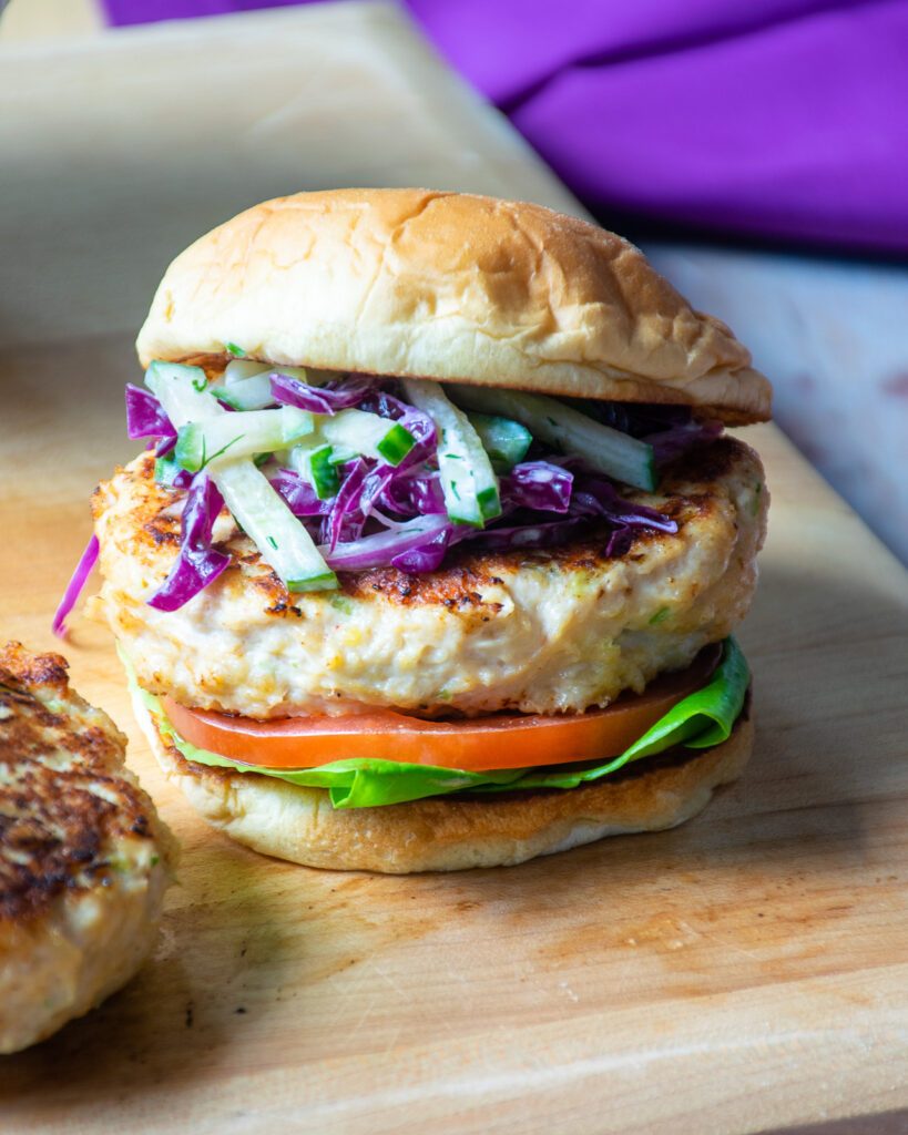 A chicken burger with cucumber slaw on a wooden cutting board with a purple napkin in the background.