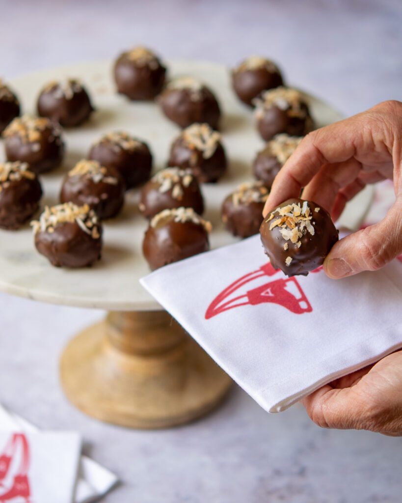 A hand holding a chocolate toasted coconut ball with a cake plate of more in the background.