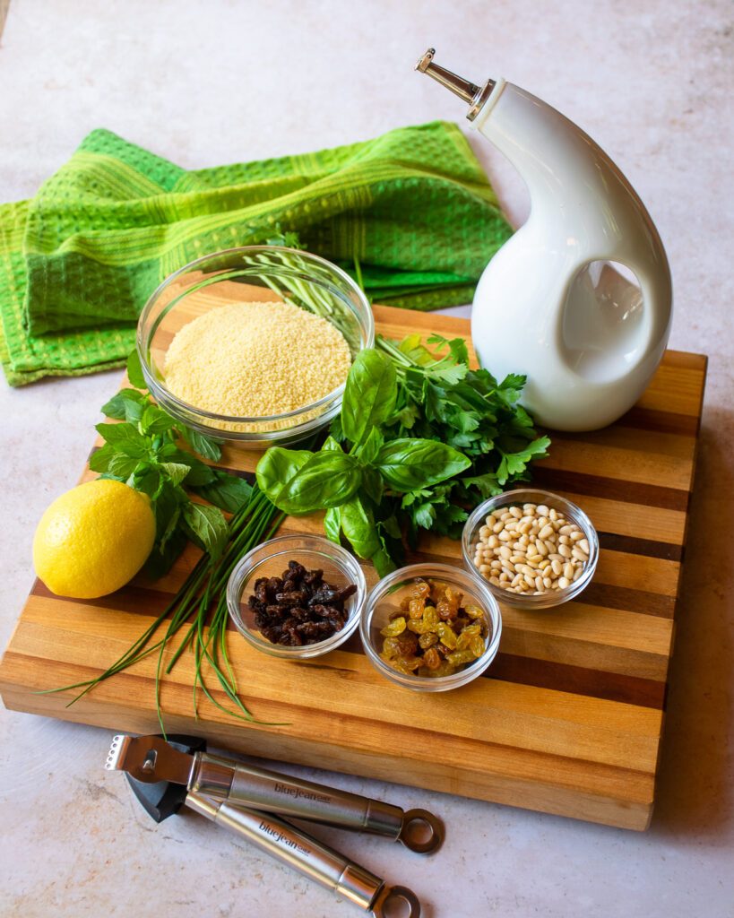 Ingredients on a cutting board on a counter - raisins, pinenuts, herbs, lemon, a bowl of couscous and olive oil - with a green kitchen towel in the background.