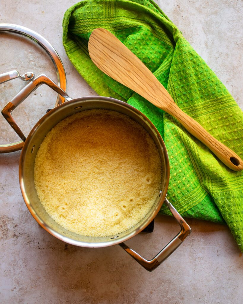 Looking down into a casserole pan with cooked couscous in it, next to a green kitchen towel and a wooden spoon.