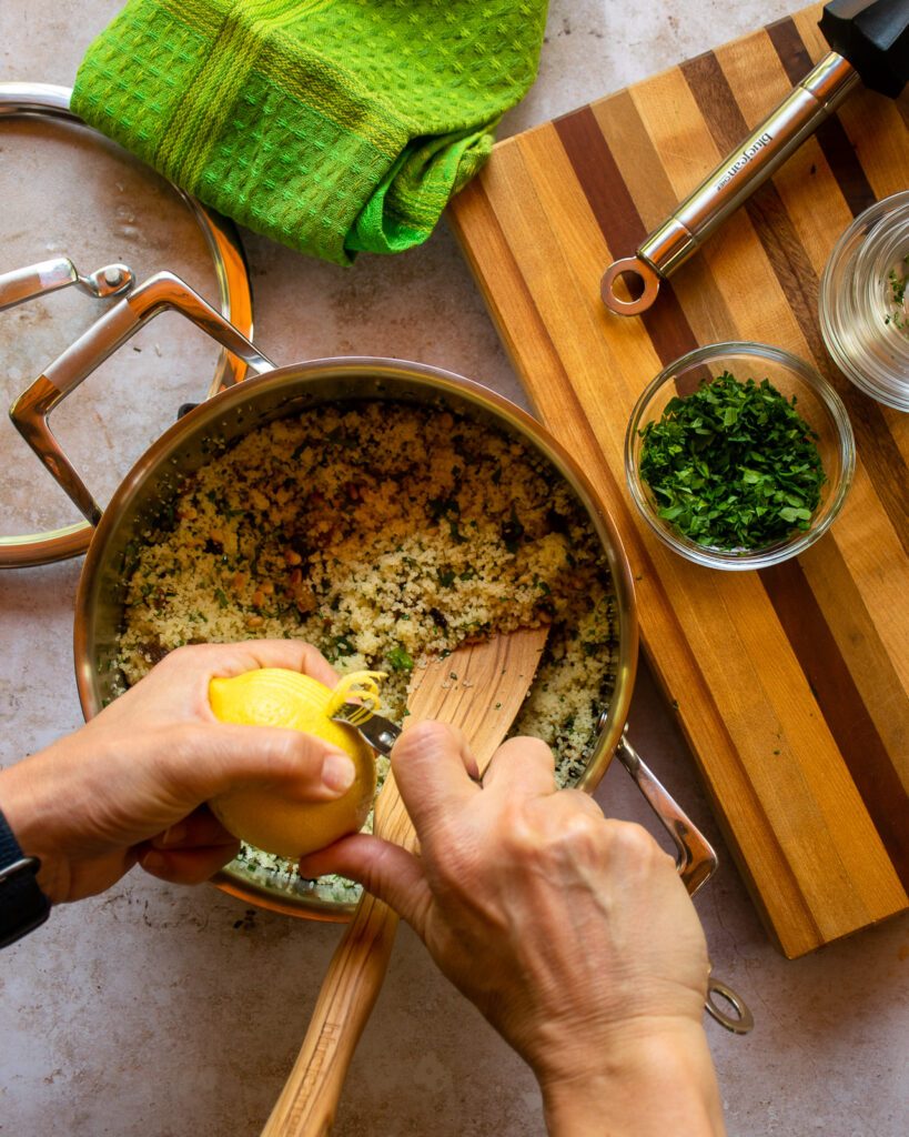 Hands using a zester to zest lemon into a pot of couscous with herbs.