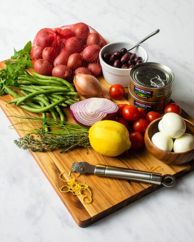 Ingredients on a wooden cutting board - red potatoes, black olives, cans of tuna, cherry tomatoes, hard boiled eggs, lemon, red onion, fresh herbs, green beans - and a citrus zester.