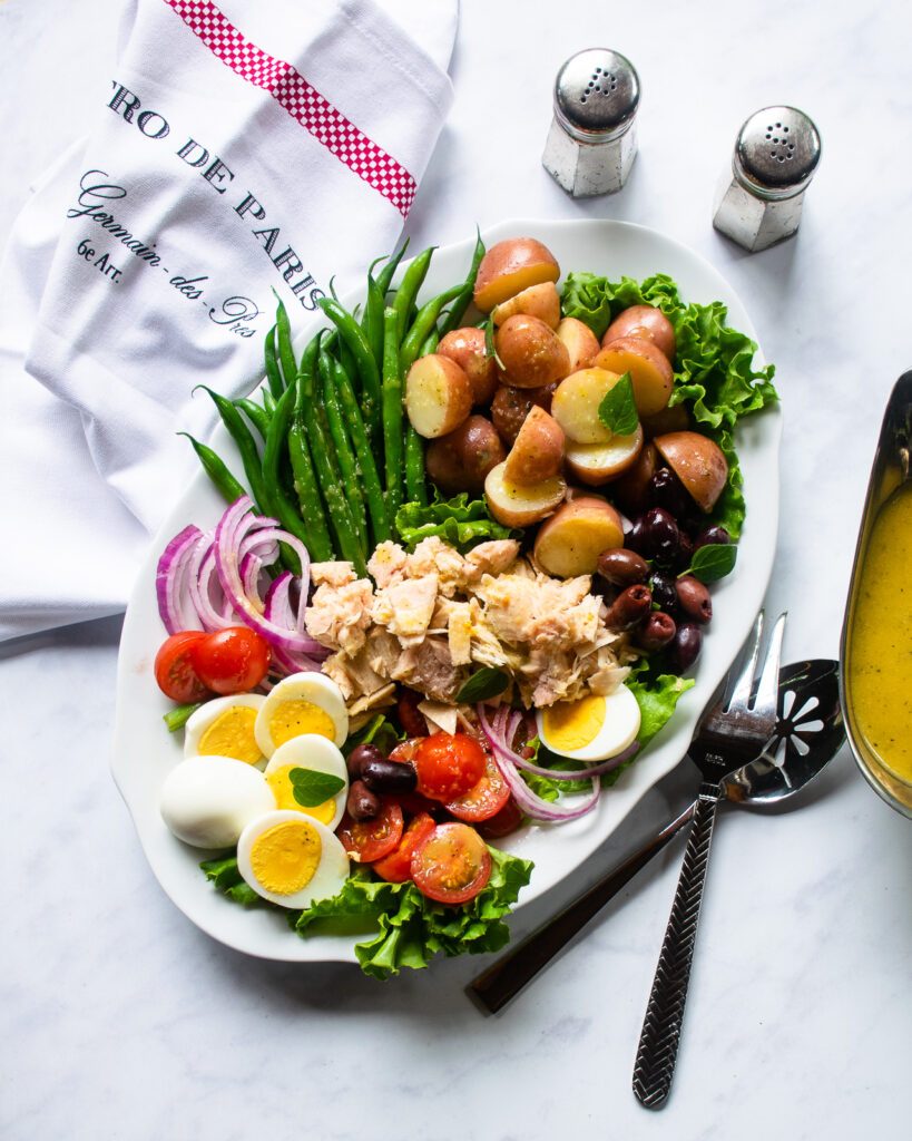 Looking down on a white oval platter of Niçoise salad with a gravy boat of dressing, salt and pepper shakers and serving utensils next to it.