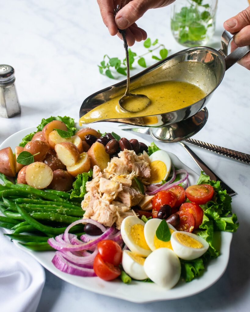 Hands dressing a Niçoise salad with salad dressing from a gravy boat.