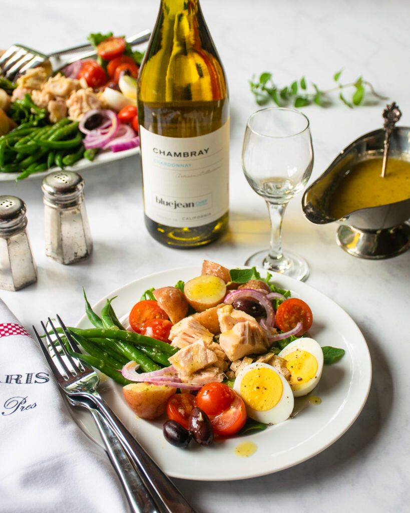A white dinner plate of Niçoise salad with utensils, a wine glass, a bottle of white wine and a platter of more salad in the background.