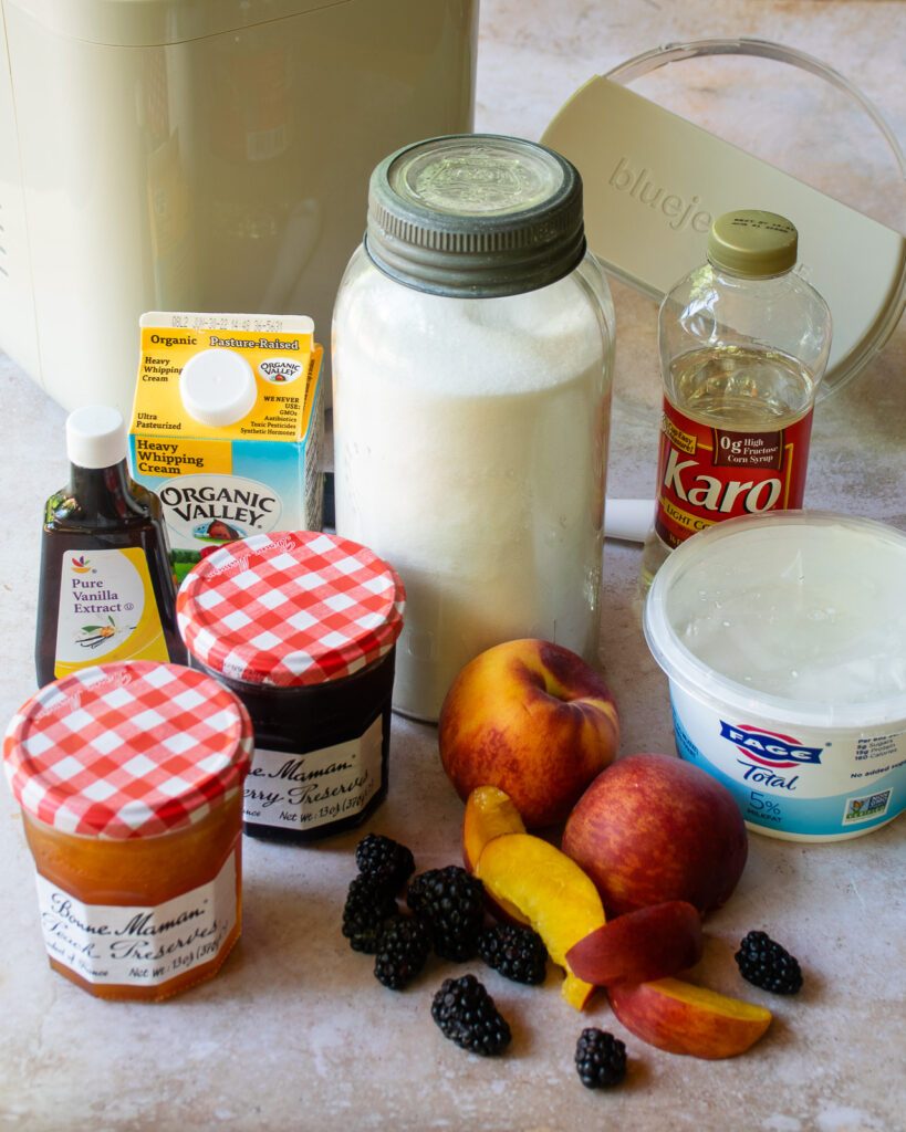 Ingredients on a countertop - yogurt, cream, sugar, corn syrup, vanilla extract, peach jam, blackberry jam.