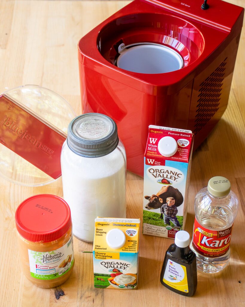 Ingredients on a butcher block counter in front of a red ice cream maker - peanut butter, cream, sugar, milk, vanilla, corn syrup.