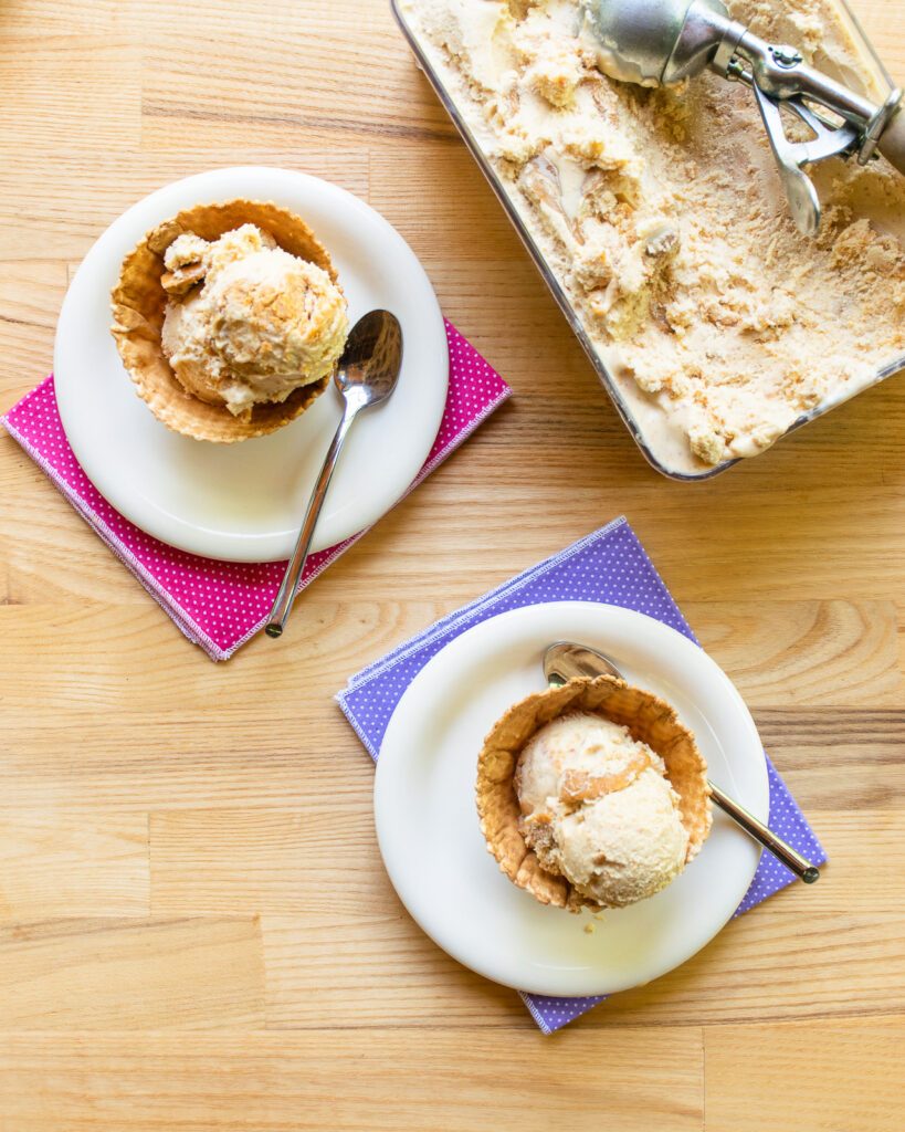 Peanut butter swirl ice cream in waffle cups on little white plates with colorful napkins underneath, next to a container of ice cream with a scoop.