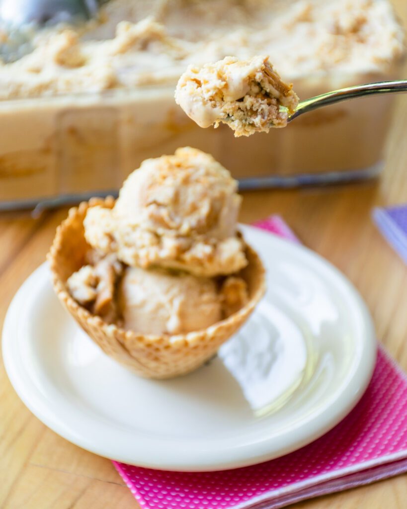 A spoon lifting up peanut butter swirl ice cream with a scoop in a waffle cup on a plate down below.