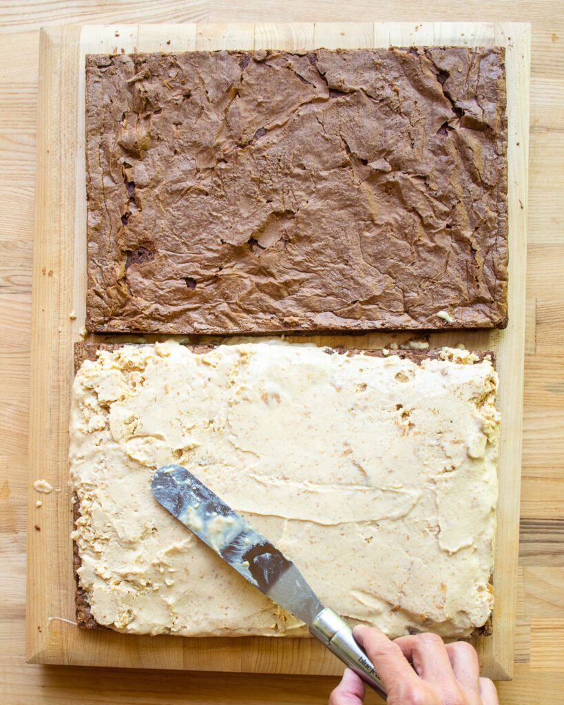A hand spreading peanut butter swirl ice cream on one half of a sheet pan brownie.