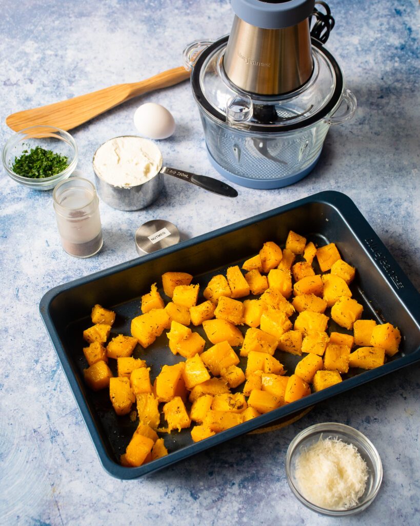 Butternut squash cubes roasted in a roasting pan with a mini chopper and some other ingredients around it.