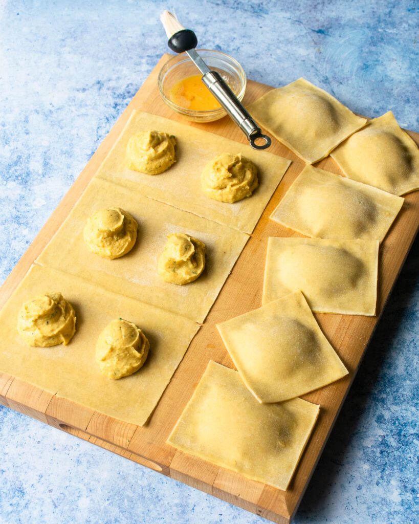 Squares of fresh pasta on a cutting board with dollops of butternut squash filling on top, next to some completed ravioli.