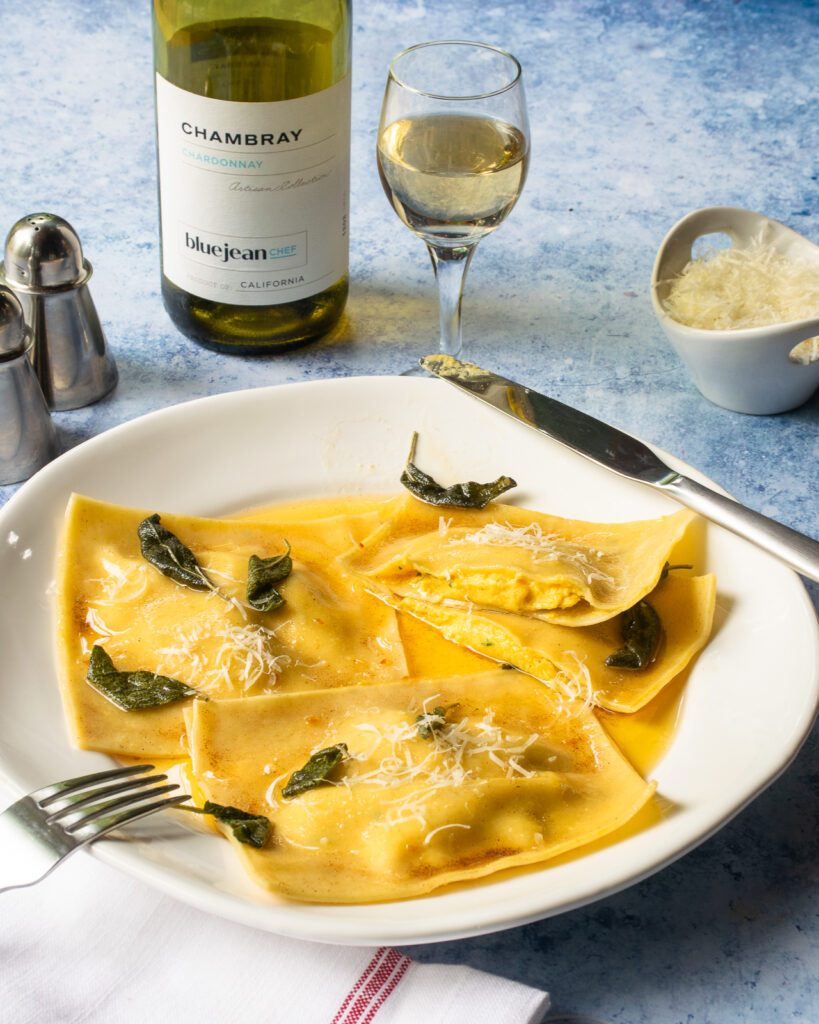 Butternut squash ravioli on a white plate with knife and fork, a glass and bottle of white wine and some grated parmesan cheese.