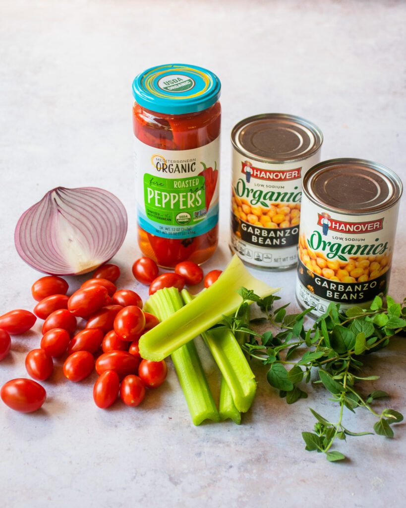 Ingredients on a counter - cherry tomatoes, celery, parsley, chickpeas, half a red onion and a jar of roasted peppers.
