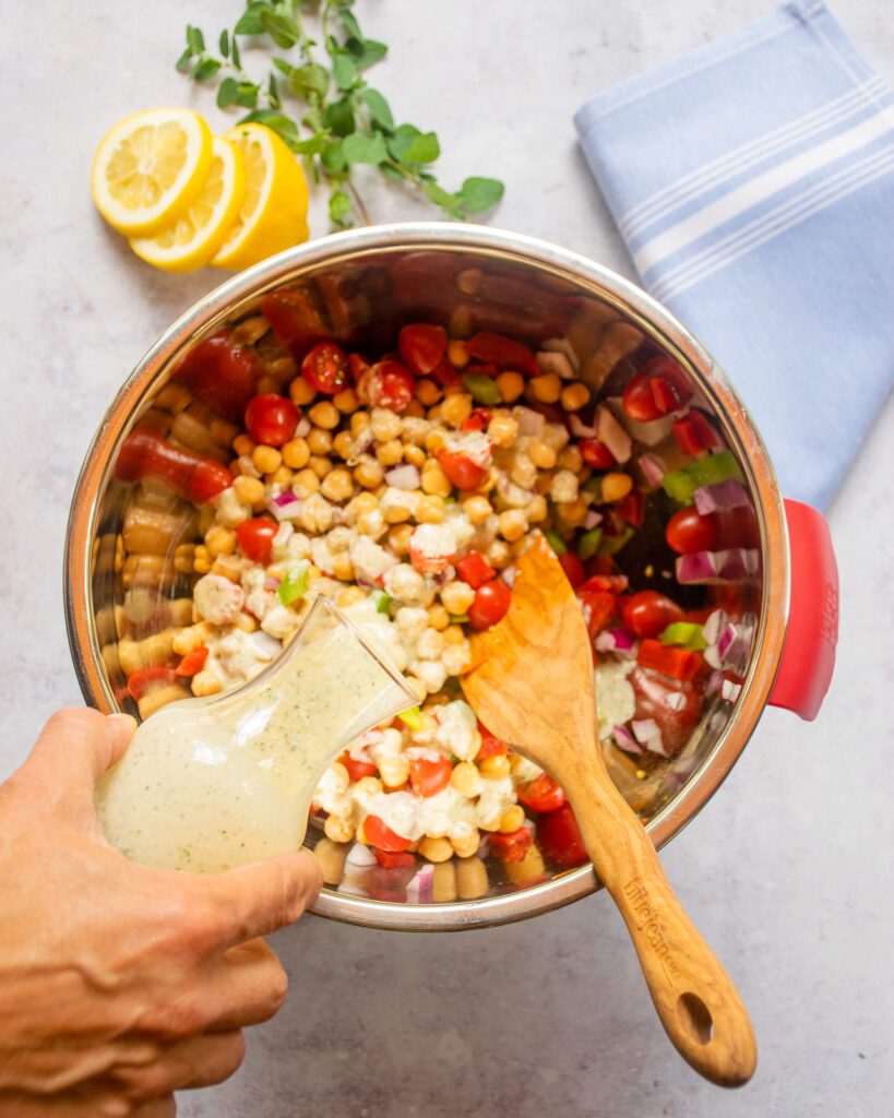 A hand pouring dressing into a bowl of chickpea salad with a wooden spoon.