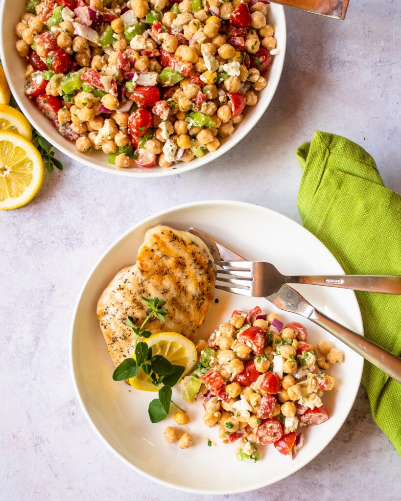 A plate with a grilled chicken breast and some chickpea salad, knife and fork and a green napkin beside it.