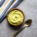 Avocado crema in a small wooden bowl with a spoon on one side and a blue and yellow striped towel on the other side.