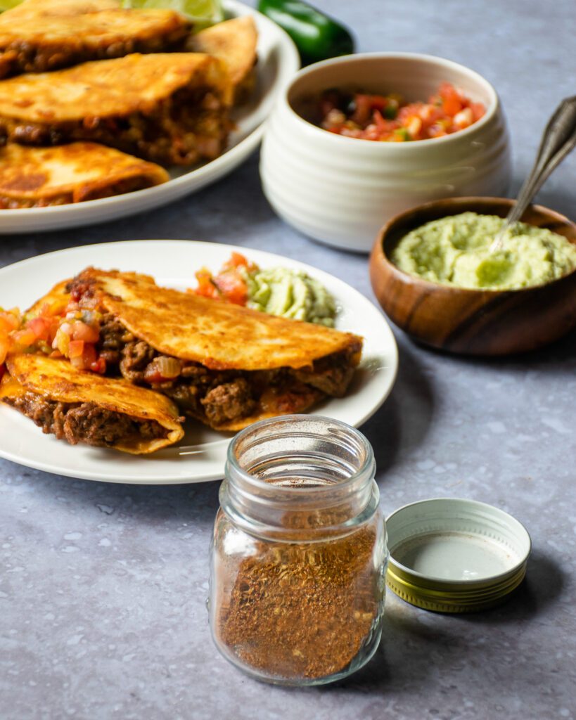 A jar of taco seasoning in front of crispy beef tacos on a white plate.