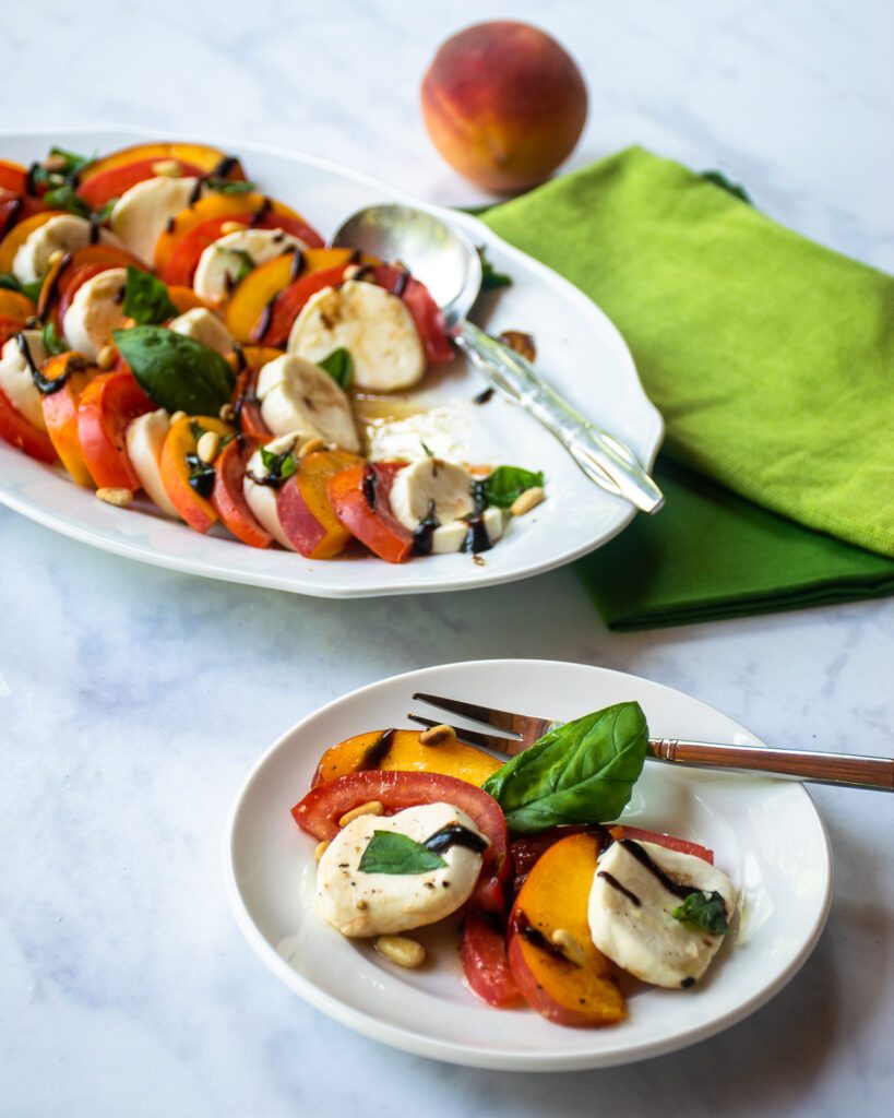 A serving of peach caprese salad on a small white plate in front of a larger oval platter.
