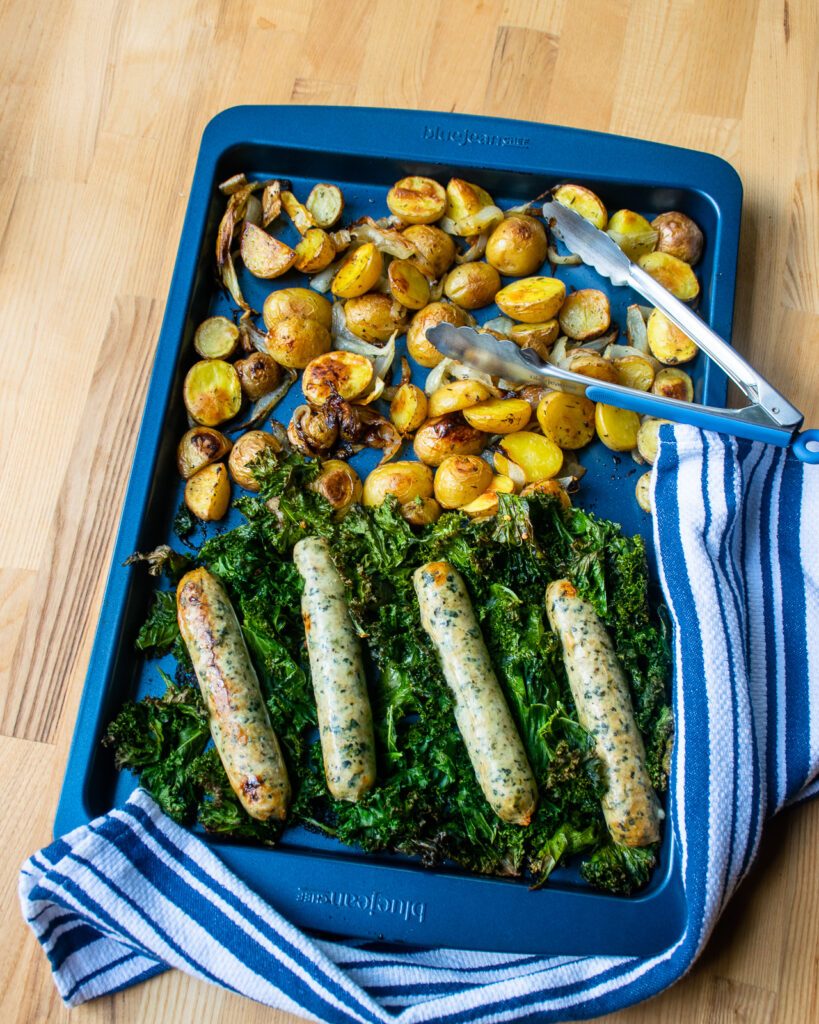Sausages, potatoes and kale on a large blue sheet pan with a pair of tongs and a blue and white striped towel.