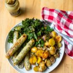A large white platter with sausages potatoes and kale next to a red and white checkered napkin and a jar of mustard.