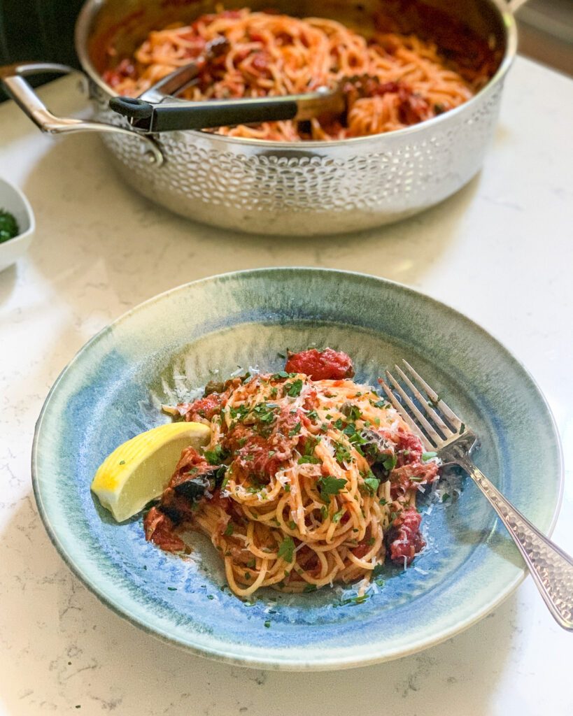 Spaghetti Tuna Puttanesca in a blue bowl with a fork and lemon wedge.