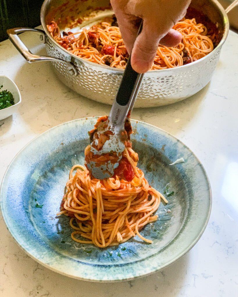 Tongs placing spaghetti with a red sauce into a blue bowl.