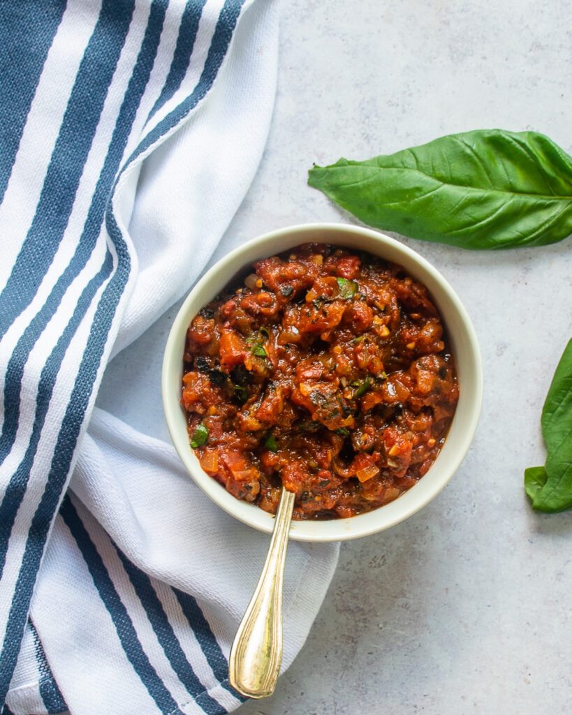 Looking down into a white bowl of tomato spread with basil leaves, a blue and white striped towel next to it.