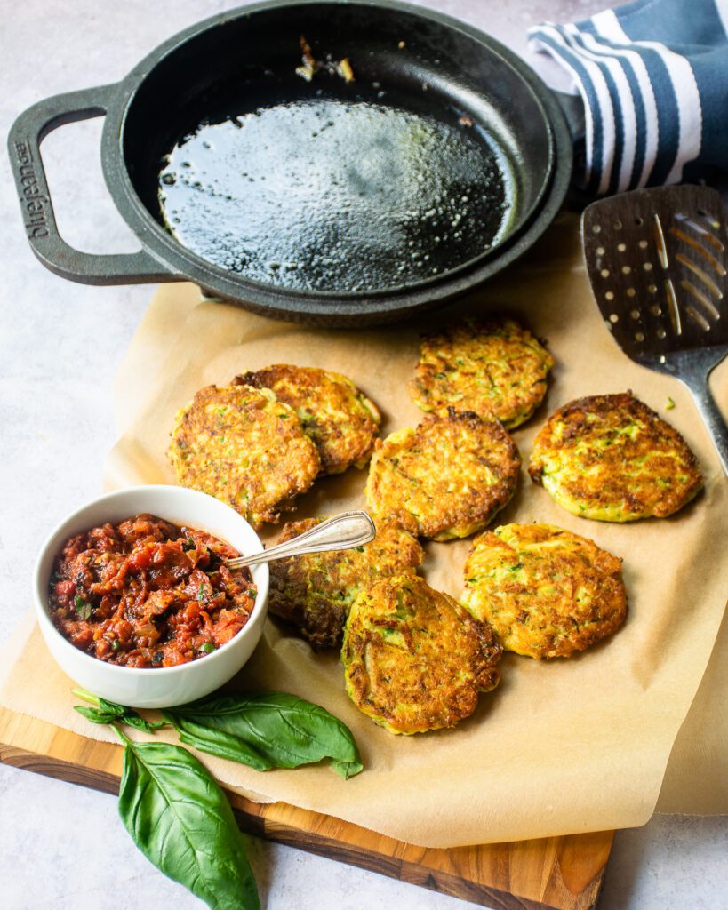 A board with zucchini fritters, fire-roasted tomato spread and a cast iron skillet in the background.