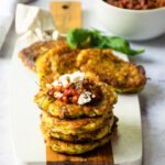A stack of zucchini fritters on a board with more in the background and a bowl of tomato spread.