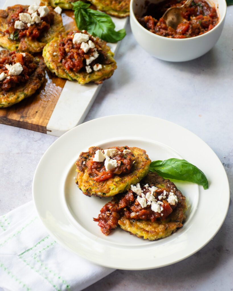 A white plate with two zucchini fritters, fire-roasted tomato spread and ricotta with more in the background.