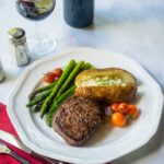 A white plate with filet mignon, baked potato and asparagus with a place setting and wine.