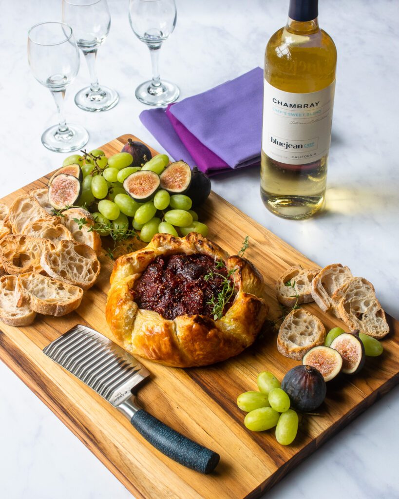 Baked Brie with Fig jam on a serving board with crostini, grapes, raw figs and a cheese knife, with wine and napkins next to it.