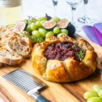 Baked brie with fig jam on a serving board with crostini, grapes and figs.