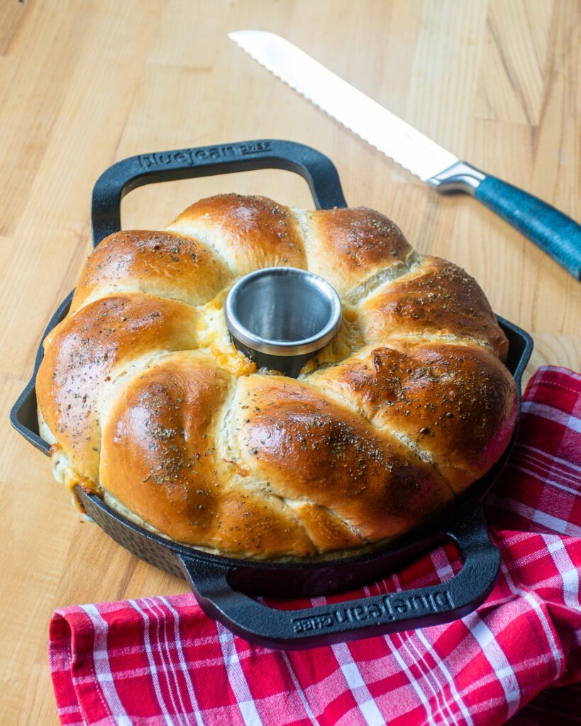 A cheese filled bread ring in a cast iron pan with a red kitchen towel and a bread knife nearby.